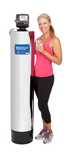 A fit older woman holding a glass of water stands next to a Waterboy 2000 SXT